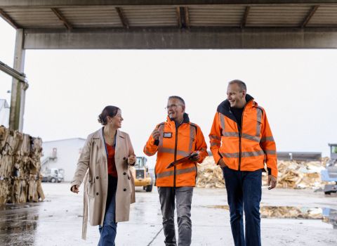 Drei freundliche Personen aus dem Weber Team, laufend auf dem Betriebshof in einer offenen Halle. Im Hintergrund sortiertes Altholz und gepresste Altpapier-Ballen.