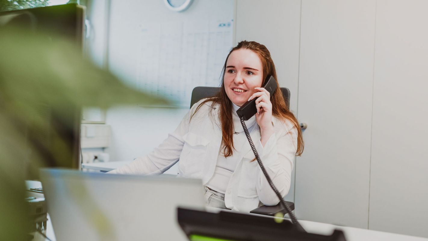Junge Ansprechpartnerin von Weber spricht am Telefon vor einem Rechner. Im Vordergrund eine unscharfe Pflanze, im Hintergrund eine helle Bürougebung.