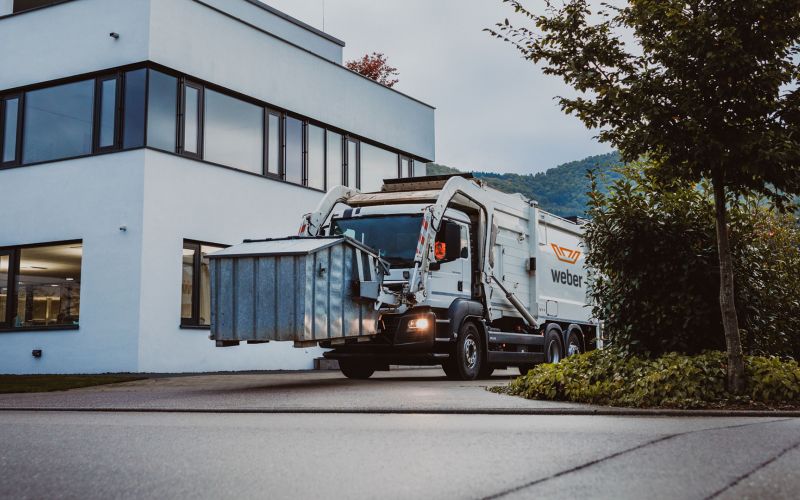 Frontlader von Weber hebt Umleerbehälter vor modernem Bürogebäude in grüner Umgebung an.