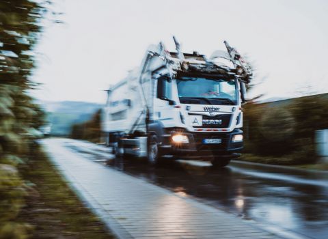 Bild mit Bewegungsunschärfe: Weißer LKW Frontlader von Weber fährt schnell über eine Landstraße im Schwarzwald. Regnerische Stimmung, Reflexionen auf der nassen Straße.