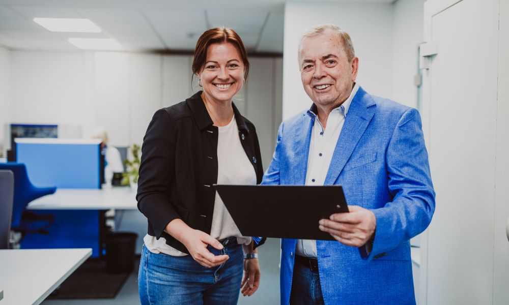 Die Geschäftsleitung (eine Frau und ihr Vater) stehen lachend im Büro, prüfen Dokumente, die der Vater in der Hand hält.
