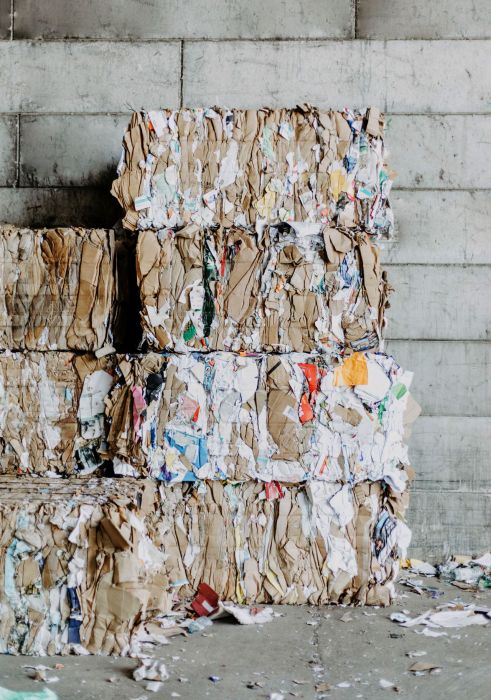 Acht übereinander gestapelte gepresste Ballen aus Altkarton auf vor einer Betonwand. Braune und bunte Kartonagen.