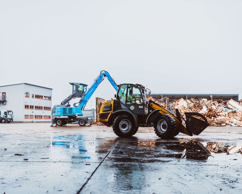 Radlader fährt auf Webers Betriebshof, im Hintergrund sortiertes Altholz und weitere Fahrzeuge.